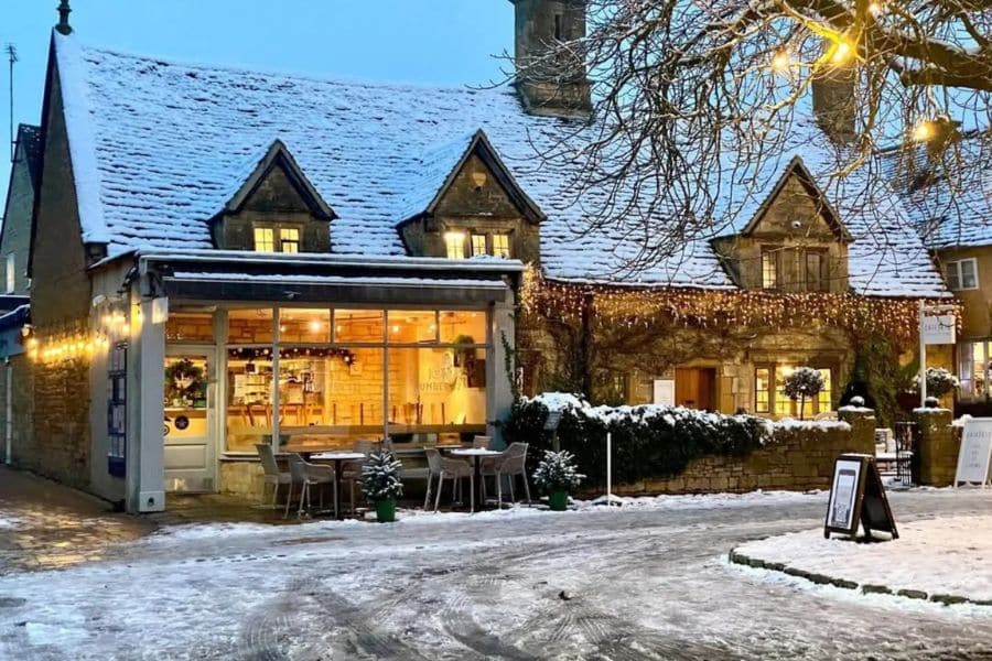 A snow-dusted tearoom in the Cotswolds glows warmly from inside, with fairy lights strung along the roof and a tree lit outside. Quaint villages like this are perfect places to visit at Christmas in the UK for a quiet, cozy getaway.