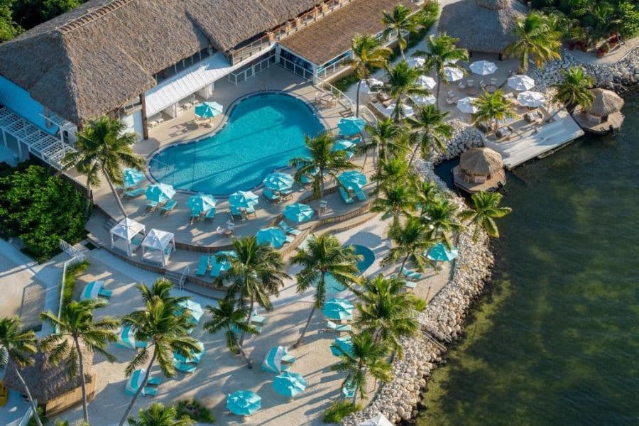 A heart-shaped pool is surrounded by turquoise umbrellas and palm trees at a waterfront resort, showcasing beachfront luxury at All-Inclusive Resorts in the US.