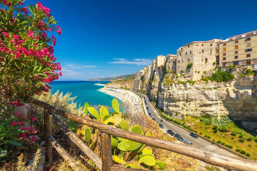 Colorful beachside buildings perched on cliffs overlook a sweeping sandy shore and clear blue sea, framed by blooming flowers and cactus plants. A picturesque escape from cold weather and a great way to escape the winter blues in January.