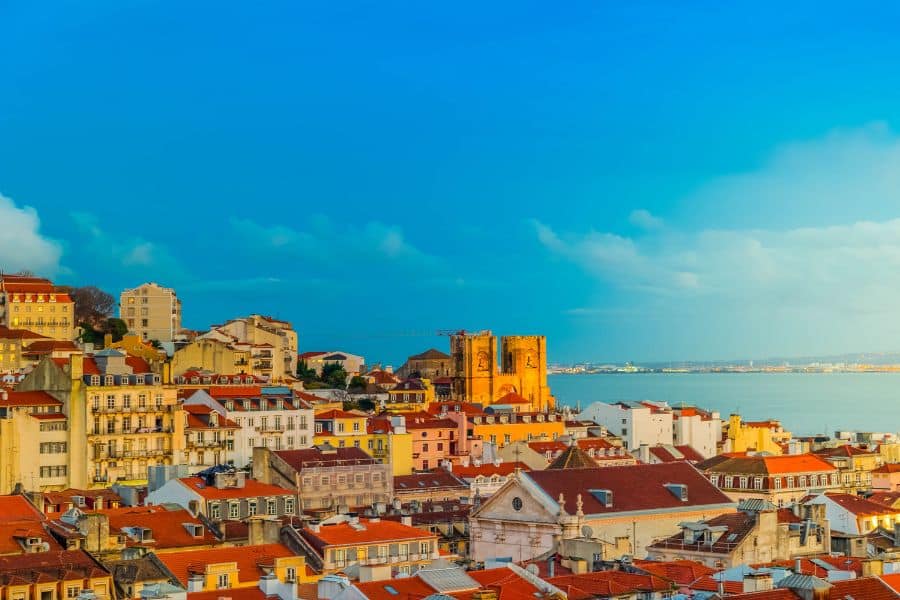 Red-tiled rooftops of Lisbon glow in warm sunlight with the Tagus River in the background and a golden sky above. A vibrant city break to escape the winter blues in January with culture, color, and coastal charm.