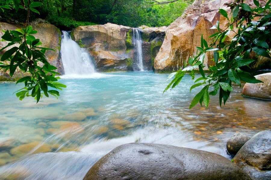 Twin waterfalls cascade into a turquoise pool surrounded by lush greenery, one of the exotic shore excursions available when you Live on a Cruise Ship.