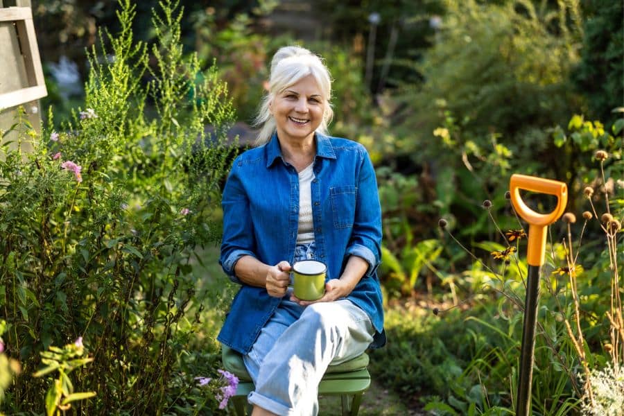 Smiling senior woman in a denim shirt holding a mug while sitting in her backyard garden, enjoying a break and showing how gardening is the #1 hobby for retirees who love nature and peace.