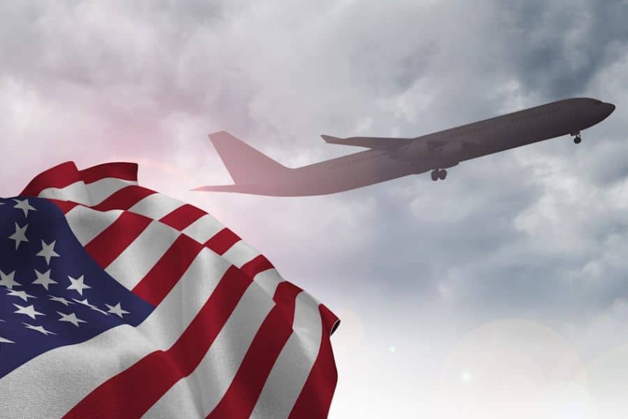 A commercial airplane flying through a cloudy sky with the American flag in the foreground, symbolizing international travel affected by the New UK Travel Rules.