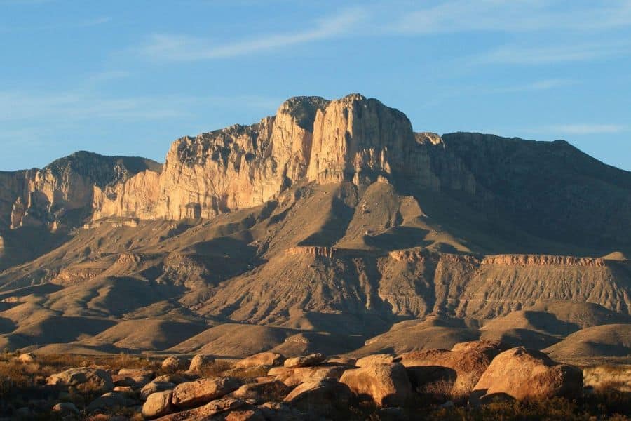 Sunlight hits the rocky face of a desert mountain under a blue sky, showcasing one of the striking yet increasingly Overcrowded US National Parks.