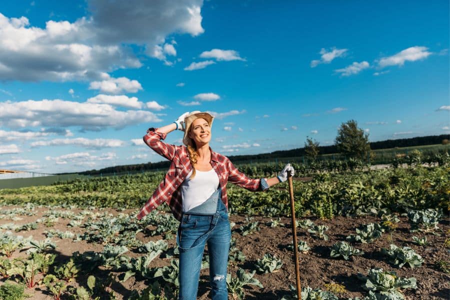 A woman standing proudly in a large vegetable garden on a sunny day, symbolizing fulfillment and independence as gardening is the #1 hobby for retirees.