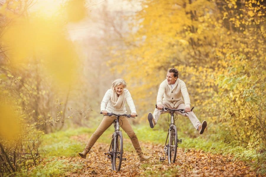 An older couple joyfully rides bicycles through a forest path covered in autumn leaves. Both are wearing cozy sweaters, with the man playfully lifting his legs off the pedals as they laugh together amidst vibrant yellow foliage.