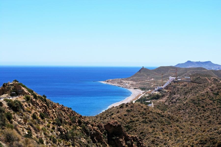 A winding coastal road hugs arid hills overlooking a crescent-shaped bay and deep blue sea under a cloudless sky. A quiet Mediterranean getaway to escape the winter blues in January away from the crowds.