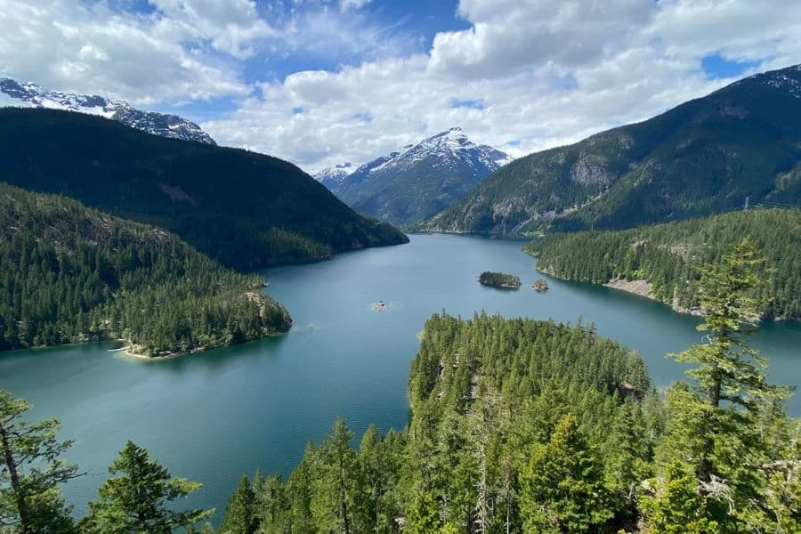 A wide mountain lake snakes through tree-covered hills under a partly cloudy sky, offering a glimpse into the landscape pressure from Overcrowded US National Parks.
