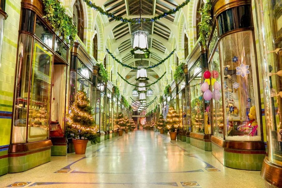 A beautifully decorated Victorian shopping arcade with garlands, mini lit Christmas trees, and large pendant lights overhead. Elegant indoor spaces like this are beloved places to visit at Christmas in the UK for festive shopping.