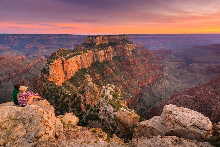 Two hikers sit at the edge of the Grand Canyon at sunset, gazing over layered cliffs and deep valleys, a serene moment in one of America’s Best-Kept Hiking Secrets beyond the crowds.