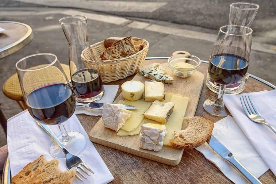 A wooden café table set with a variety of cheeses, crusty bread, and red wine, captured from a Paris sidewalk. Balanced meals with moderate indulgences help explain why the French are living longer than the Americans.