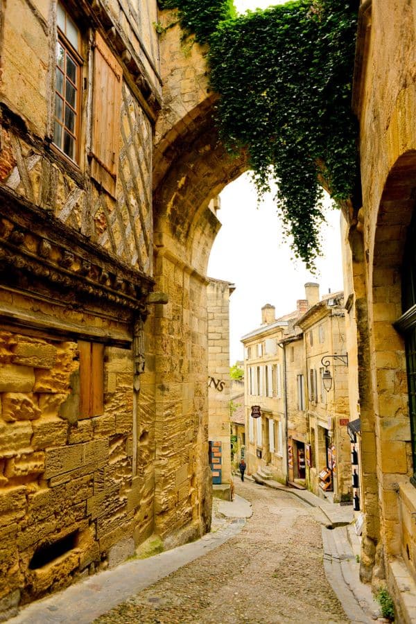 A narrow cobblestone alley under a stone archway in Saint-Émilion, France, lined with ivy-covered and timber-framed medieval buildings, exemplifies charming Medieval Places to Visit in Europe.