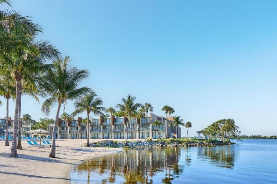 A modern beachfront resort with rows of balconies overlooks calm water and is surrounded by palm trees and sandy shoreline, showcasing tropical tranquility at one of the best All-Inclusive Resorts in the US.