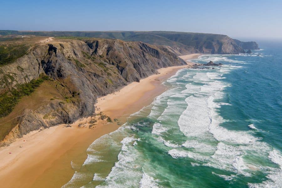 Tall cliffs tower over a vast sandy beach with rolling waves crashing along Portugal’s rugged Atlantic coast. A wild and windswept spot to escape the winter blues in January for those craving nature and solitude.