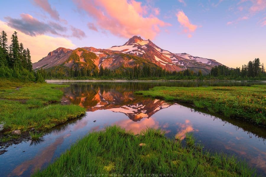 A glowing alpine sunset reflects off a clear lake surrounded by green meadows and forest, a view often chased by visitors in Overcrowded US National Parks.