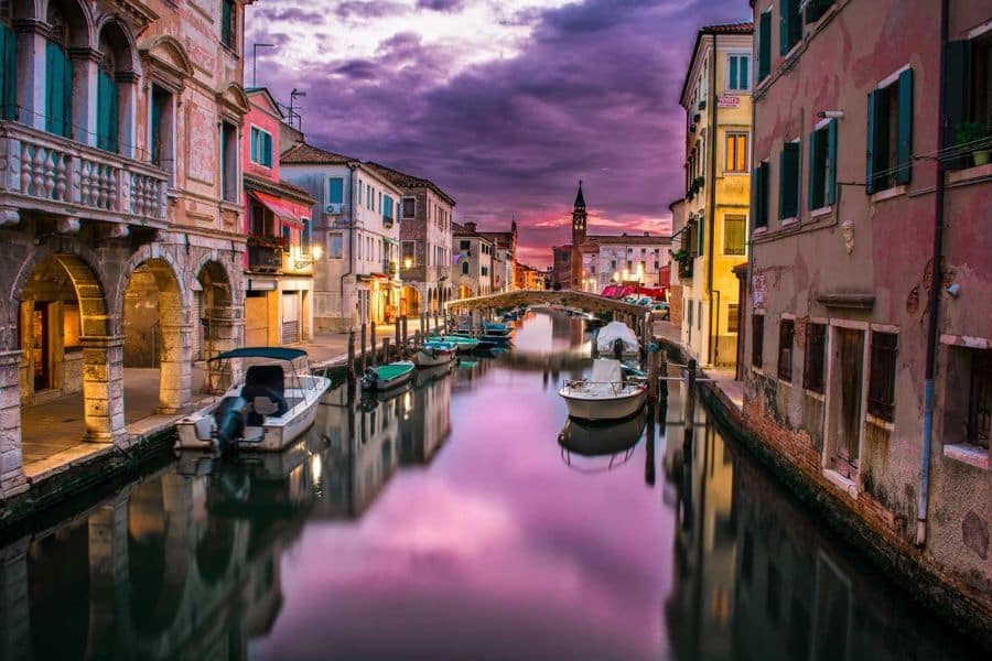 A quiet canal in Venice at sunset, lined with colorful old buildings and small boats gently floating on the water. The sky glows with dramatic purple and pink hues, reflected perfectly in the still canal beneath a stone footbridge.