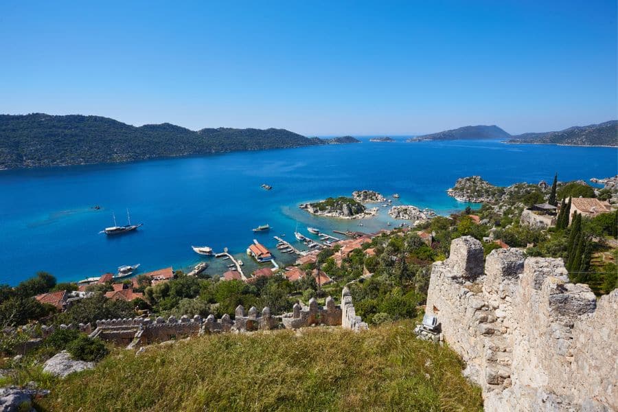 View of a serene harbor dotted with sailboats and ancient stone ruins on the Turkish coast, one of the historic stops you could visit if you choose to Live on a Cruise Ship.