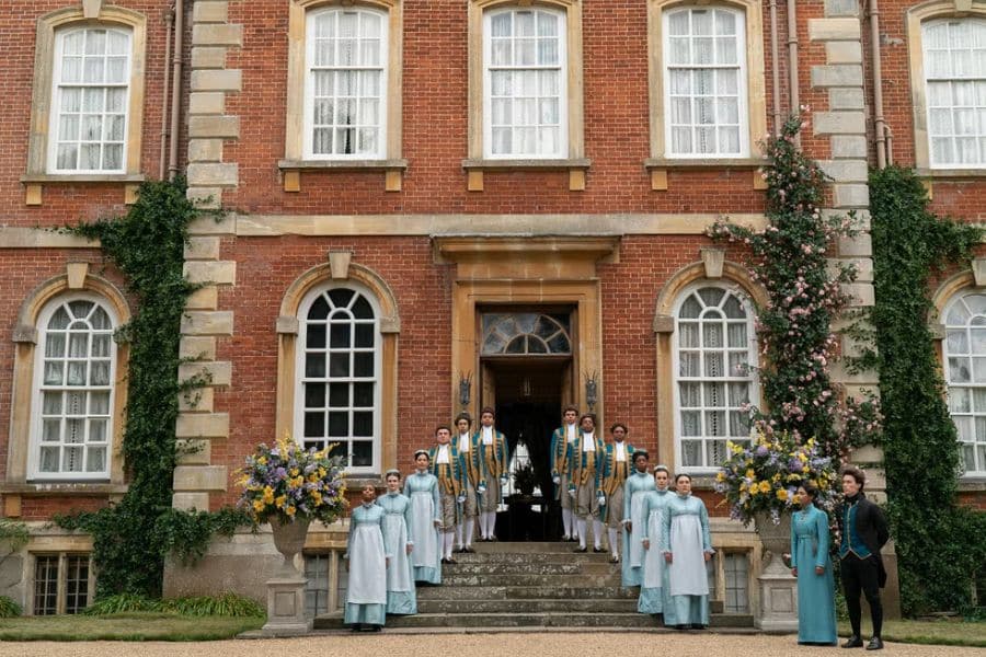 A group of formally dressed individuals in pastel Regency-era costumes stands in formation outside a grand red-brick mansion with white-trimmed windows. This filming site is one of the iconic Bridgerton locations known for its elegant exteriors.