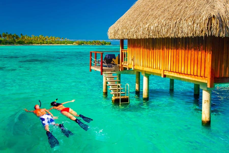 A floating bungalow on blue water with two people floating nearby with scuba gear.