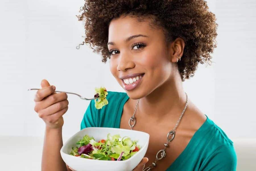A smiling woman in a teal shirt holds a forkful of leafy green salad from a white bowl. Leafy greens like these are part of the 7 Foods That Actively Fight Wrinkles due to their high vitamin content.