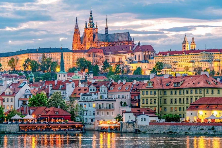 Scenic view of Prague at dusk with Prague Castle and St. Vitus Cathedral illuminated above the Vltava River, showcasing one of Europe's capital cities full of history.