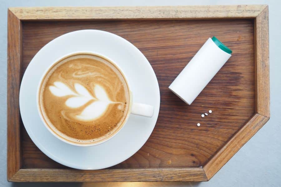 Cup of frothy coffee with sugar substitute tablets on a wooden tray, illustrating artificial sweeteners as one of the 9 common foods to avoid.