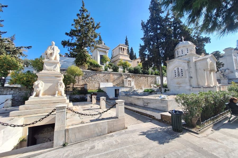 Sunlit tombs and mausoleums in the First Cemetery of Athens, Greece, surrounded by tall trees and neoclassical sculptures.