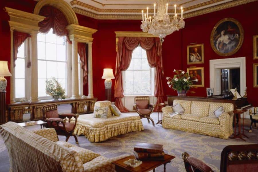 Lavish red-walled drawing room with ornate gold curtains, vintage furniture, classical paintings, and a sparkling chandelier. This interior space reflects the opulence of Bridgerton locations used to depict aristocratic homes.
