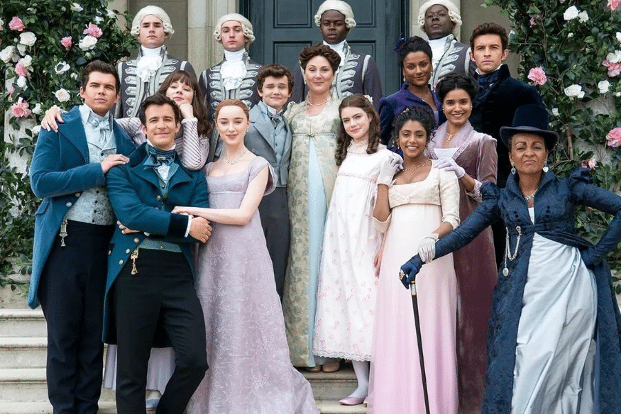 A group photo of characters from Bridgerton dressed in vibrant Regency-era costumes, standing together on stone steps decorated with roses. This ensemble cast scene is set at one of the signature Bridgerton locations used as a family estate backdrop.