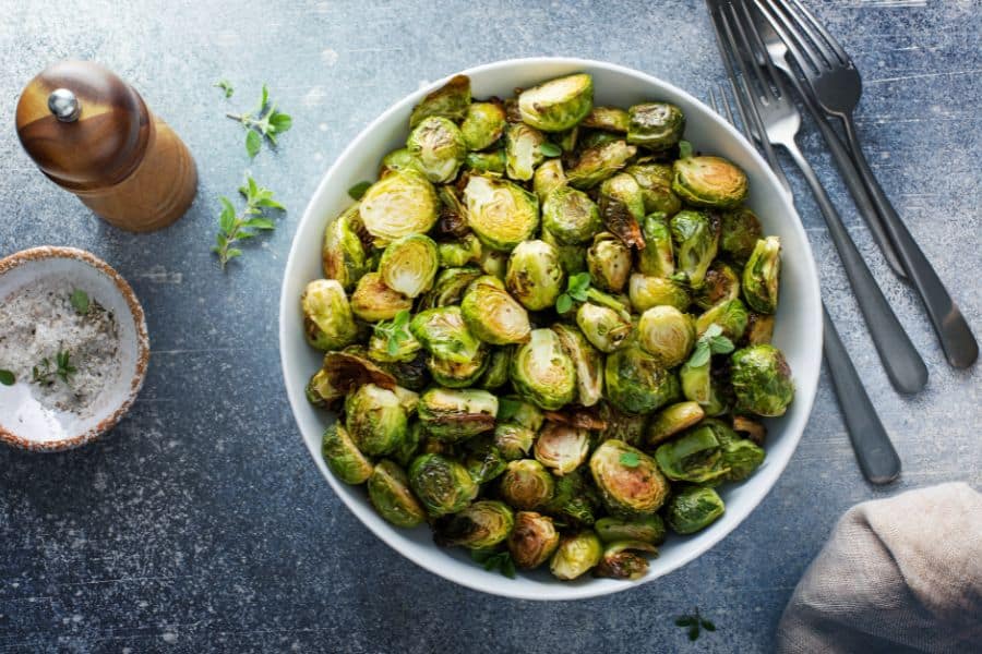 A white bowl filled with roasted Brussels sprouts, garnished with fresh herbs, sitting next to salt, pepper, and cutlery. A simple yet powerful anti-aging superfood dish.