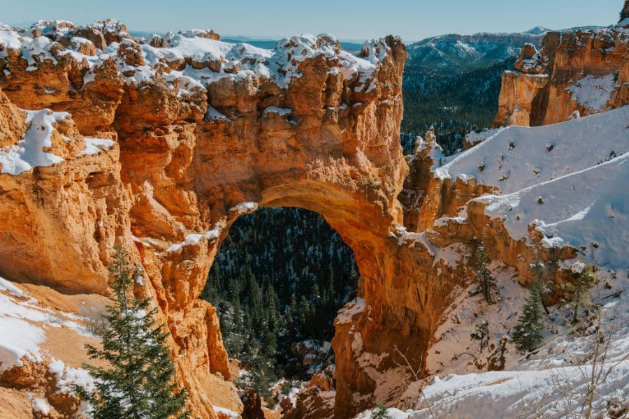 A natural sandstone arch dusted with snow frames a view of dense forest in Bryce Canyon National Park, Utah. This unique winter path highlights the diversity of hiking trails in the US.