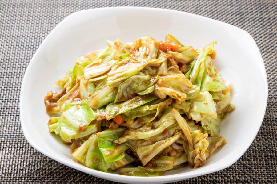 A plate of stir-fried cabbage with a light brown sauce and visible caramelized edges, served in a white bowl on a woven placemat. Cabbage is a cruciferous vegetable linked to cellular health and longevity.