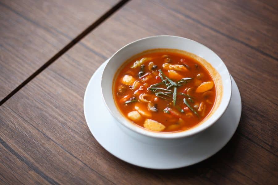Bowl of hearty vegetable soup in a red broth on a wooden table, which may be high in sodium and part of the 9 common foods to avoid.