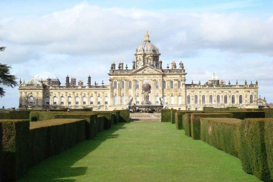 A long manicured lawn leads up to the majestic Castle Howard, with its baroque architecture and central dome under a blue sky. Castle Howard is famously featured as one of the Bridgerton locations, representing the Duke of Hastings' estate.