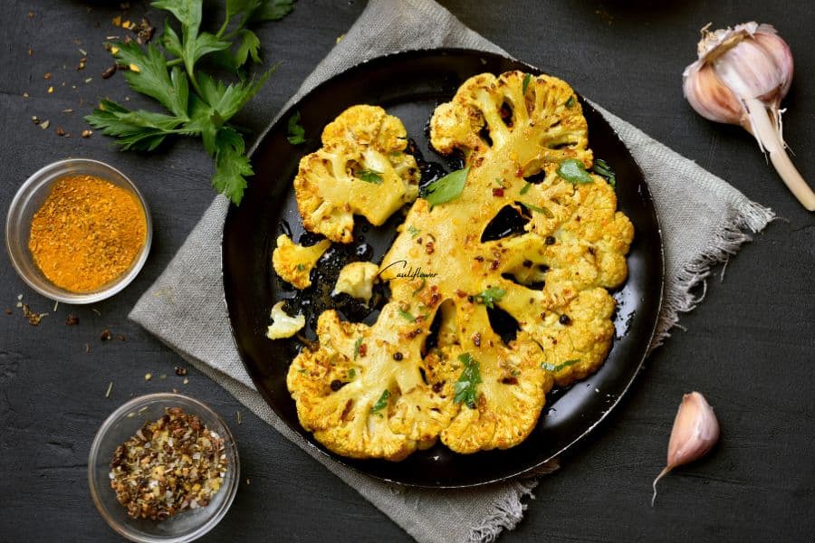 Golden roasted cauliflower steaks on a black plate, garnished with herbs and surrounded by spices and garlic cloves. Cauliflower is rich in antioxidants and supports healthy aging.