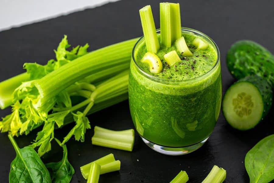 A vibrant green celery juice smoothie in a glass, surrounded by fresh celery stalks, cucumber slices, and spinach leaves. This hydrating blend includes several superfoods that help slow the aging process.