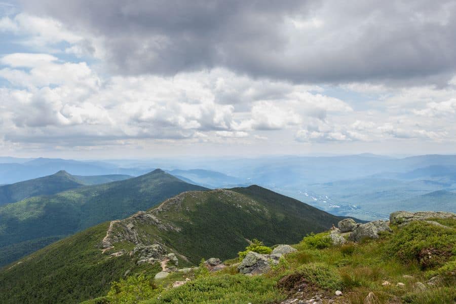 A rocky ridgeline trail winds through the White Mountains under dramatic cloud cover in New Hampshire. The Appalachian Trail is one of the most historic long-distance hiking trails in the US.