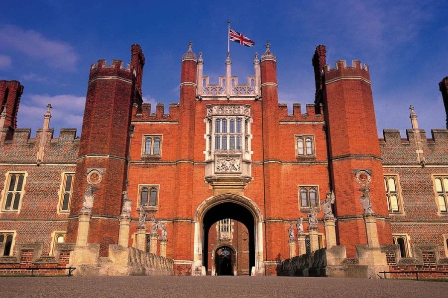 Grand entrance of Hampton Court Palace with its red Tudor brickwork, arched gate, and British flag flying overhead. This historic site stands in for several Bridgerton locations due to its period architecture.