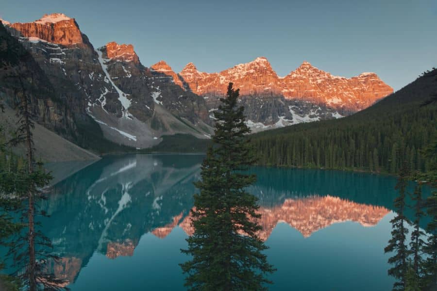 Sunrise casts a golden glow on the jagged peaks and still blue waters of Moraine Lake in Banff National Park. While located in Canada, its proximity to US border trails inspires many hiking trails in the US.