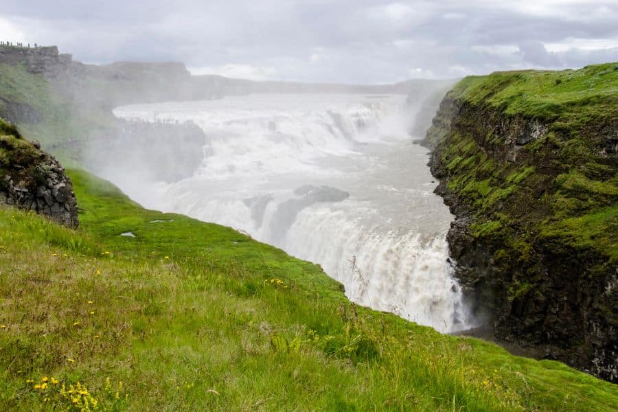 Iceland’s Gullfoss waterfall plunges between mossy cliffs, surrounded by mist and wildflowers. This natural wonder is a dramatic highlight on many travelers’ Bucket List Destinations to Visit.