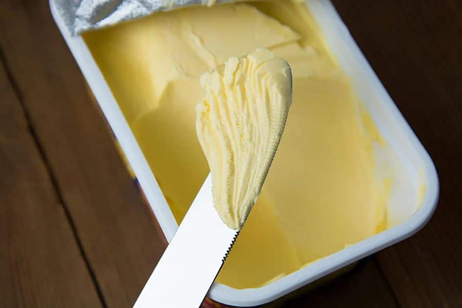 Butter knife scooping yellow margarine from a plastic container, showing processed spreads often included in the 9 common foods to avoid.