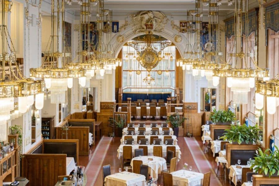 Elegant café interior with high ceilings, gold chandeliers, and art nouveau details, featuring neatly arranged tables and booth seating. This refined setting is one of the Beautiful and Historic Cafés in Europe, known for its timeless grandeur and architectural beauty.
