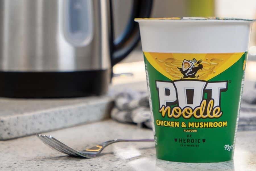 Pot Noodle Chicken & Mushroom flavor next to a kettle and fork, representing instant noodles as one of the 9 common foods to avoid.