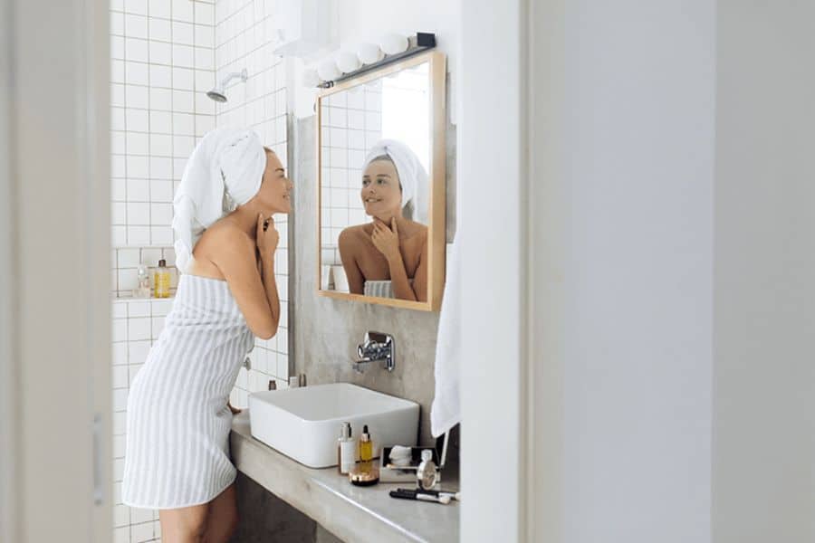 A woman wrapped in a striped towel and hair turban smiles at herself in the mirror while doing skincare in a minimalist bathroom. This self-care ritual reflects Why French Women Over 50 Look and Feel Younger.