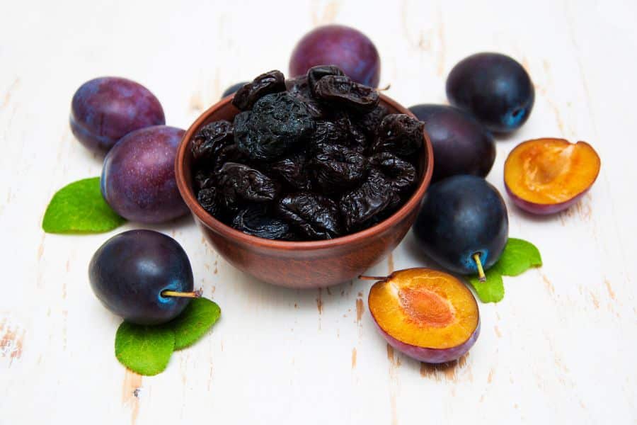 A wooden bowl filled with prunes surrounded by fresh purple plums, some whole and some halved. Prunes are high in antioxidants and fiber, and are recognized as superfoods that help slow the aging process.