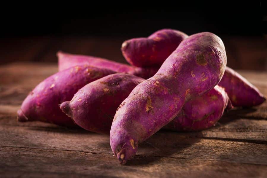 A bunch of vibrant purple sweet potatoes with rough skins laid on a wooden table. These nutrient-rich tubers are one of the 7 Foods That Actively Fight Wrinkles thanks to their high antioxidant levels.
