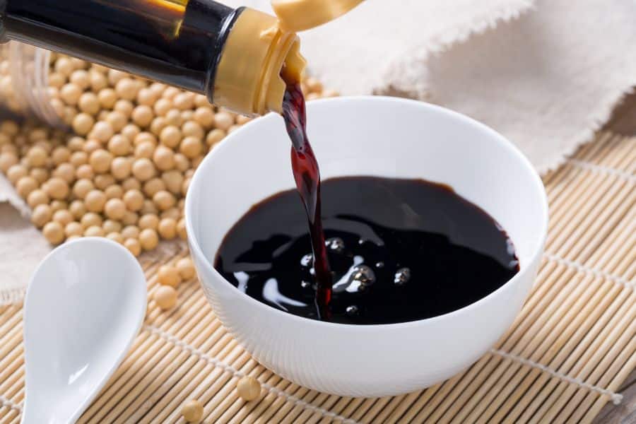 Soy sauce being poured into a small white bowl with soybeans in the background, illustrating high-sodium condiments in the 9 common foods to avoid.