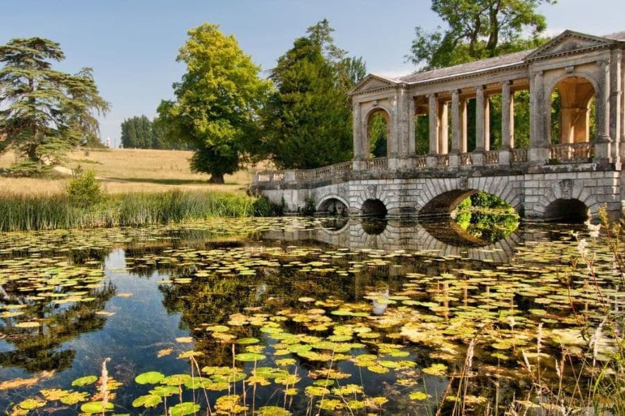 Arched stone bridge over a lily-filled pond surrounded by tall grass and trees, reflecting calm waters and romantic scenery. This picturesque spot is used among Bridgerton locations for its idyllic natural beauty.