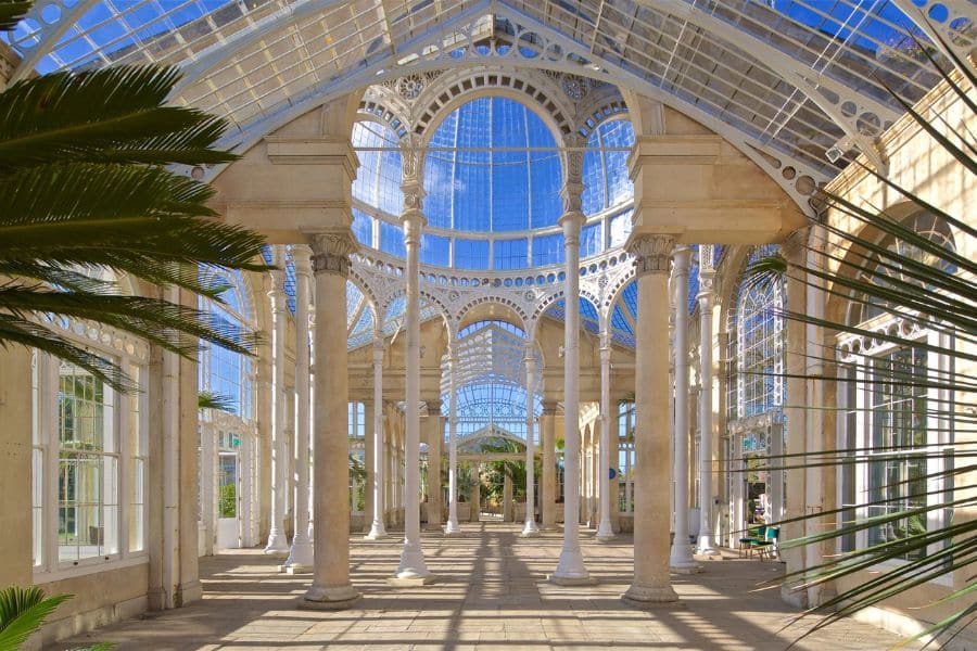 Interior of a grand glass conservatory with tall white columns, arched windows, and intricate ironwork ceiling. This light-filled space is one of the Bridgerton locations used for elegant indoor events or garden scenes.