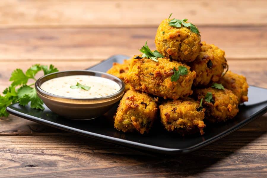 A black plate stacked with crispy golden millet fritters, garnished with cilantro and served with a creamy dipping sauce. Millet is a nutrient-dense grain often included in diets focused on longevity.